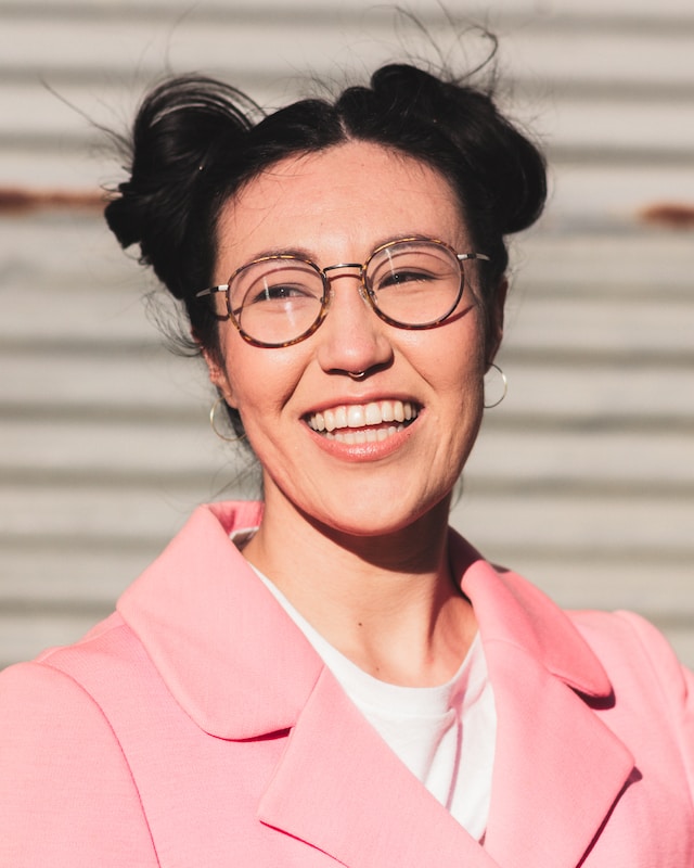 Headshot of woman with black hair tied in two buns, wearing brown glasses, medium-sized hoop earrings and a pink suit, looking towards the left and smiling.