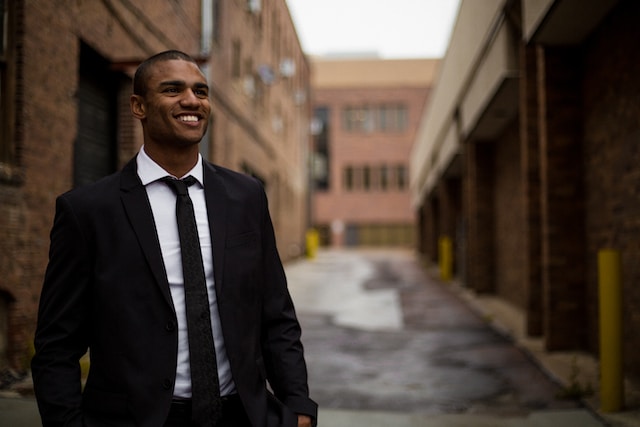 A young businessman dressed in a suit walks down an alleyway, smiling and looking upwards